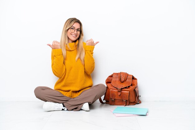 Femme étudiante uruguayenne assise sur le sol isolé sur fond blanc avec le geste du pouce levé et souriant