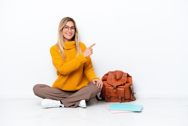 Femme étudiante uruguayenne assise sur le sol isolée sur fond blanc pointant vers le côté pour présenter un produit