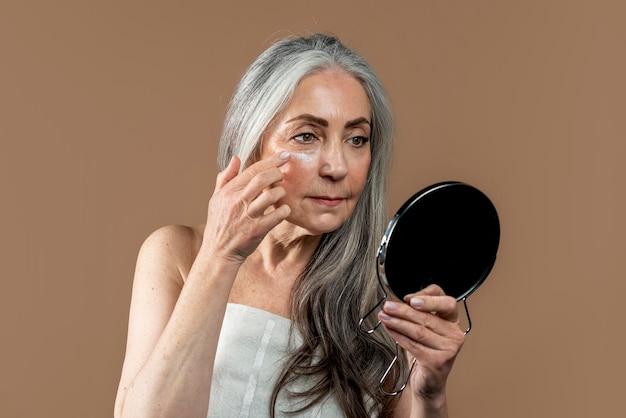 Une femme européenne âgée sérieuse aux cheveux gris dans une serviette applique une crème hydratante sur le visage regarde dans le miroir