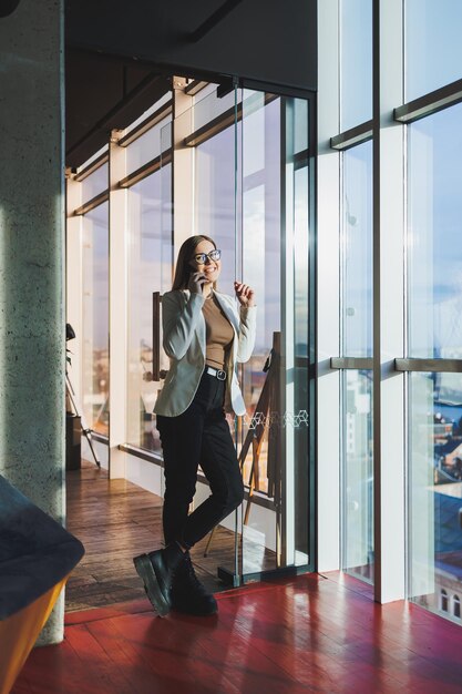 Une femme européenne aux cheveux blonds dans une veste blanche et des lunettes parle au téléphone tout en se tenant dans un bureau spacieux avec de grandes fenêtres