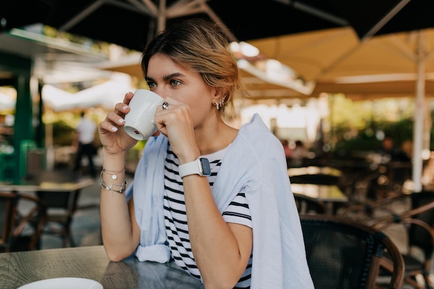 Une femme européenne aux cheveux clairs portant un t-shirt rayé et une chemise bleue boit du café le matin dans un café en plein air le matin