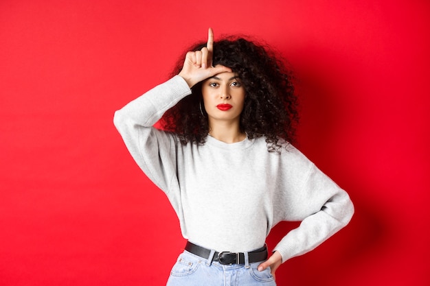 Femme européenne confiante aux cheveux bouclés, se moquant de l'équipe perdue, montrant un geste de perdant et regardant la caméra sans impression, debout dans des vêtements décontractés sur fond rouge.