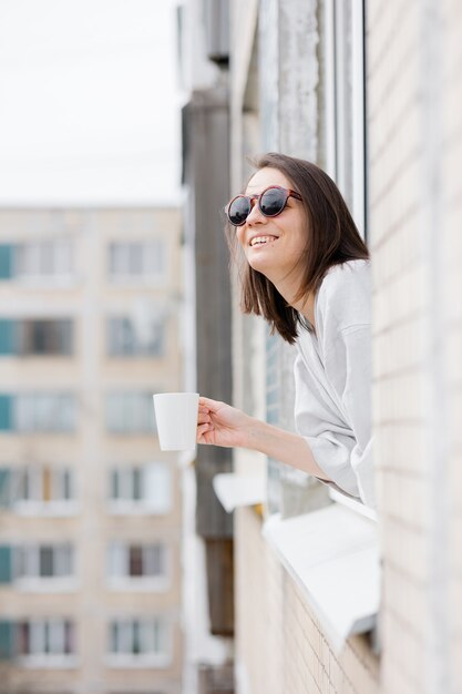 Femme européenne dans des lunettes de soleil et avec une tasse de café ou de thé regardant par la fenêtre et souriant