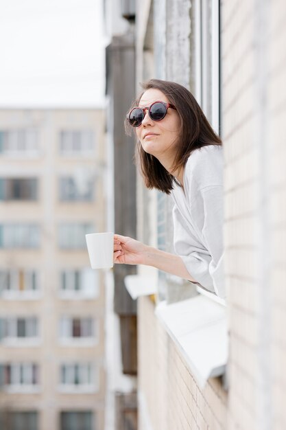 Femme européenne dans des lunettes de soleil et avec une tasse de café ou de thé regardant par la fenêtre et souriant