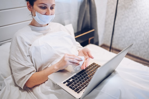 Femme européenne dans un masque facial dans la chambre pendant l'isolement du coronavirus, nettoyage de la quarantaine à la maison, ordinateur portable à la main, désinfectant pour les mains, à l'aide de coton avec de l'alcool pour éviter de contaminer avec le virus Corona.