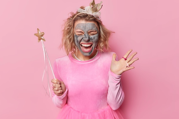 Une femme européenne émotionnelle applique un masque d'argile pour le rajeunissement subit des traitements anti-âge tient une baguette magique prétend être une princesse vêtue de vêtements de fête isolés sur fond rose