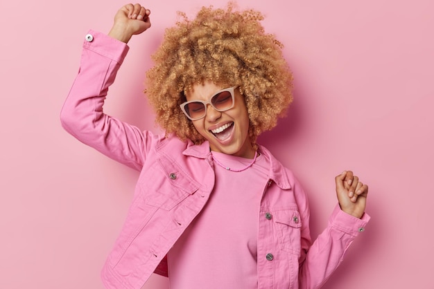 Une femme européenne gaie et insouciante aux cheveux blonds bouclés s'amuse à danser et à bouger au rythme de la musique rit joyeusement porte une veste à la mode des lunettes de soleil à la mode isolées sur fond rose