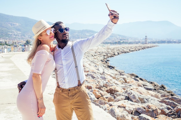 Femme européenne heureuse souriante en chapeau de paille et robe rose sexy faisant un portrait de selfie sur l'appareil photo d'un smartphone avec un homme ethnique afro-américain près de la plage d'été avec vue sur les rochers dans la station balnéaire tropicale de Turquie