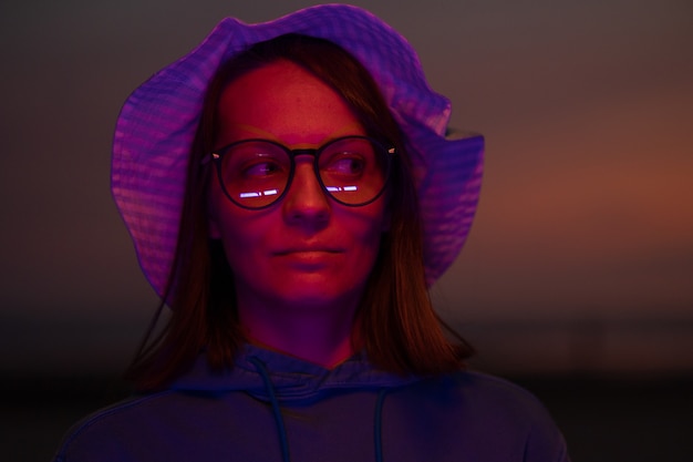 Une femme européenne illuminée par un néon dans un chapeau sur fond de ciel coucher de soleil