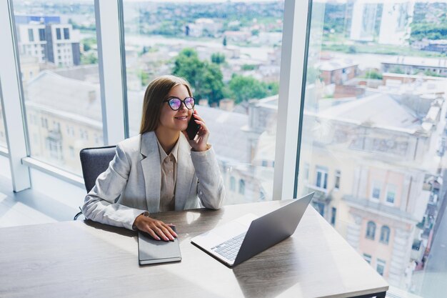 Une femme européenne joyeuse aux cheveux blonds dans des lunettes dans des vêtements décontractés élégants est assise à une table avec un ordinateur portable faisant de la paperasse et parlant au téléphone