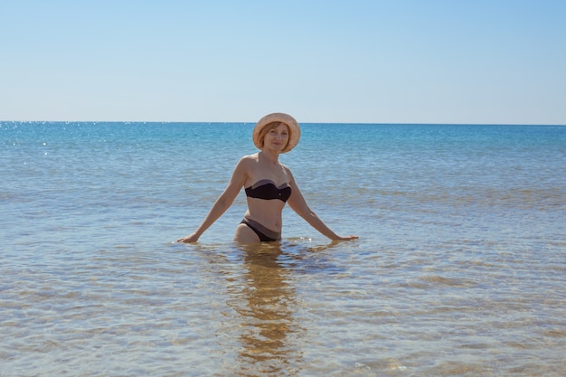 Femme européenne en maillot de bain et chapeau jusqu'à la taille dans la mer sourit et regarde la caméra. Vacances au bord de la mer.