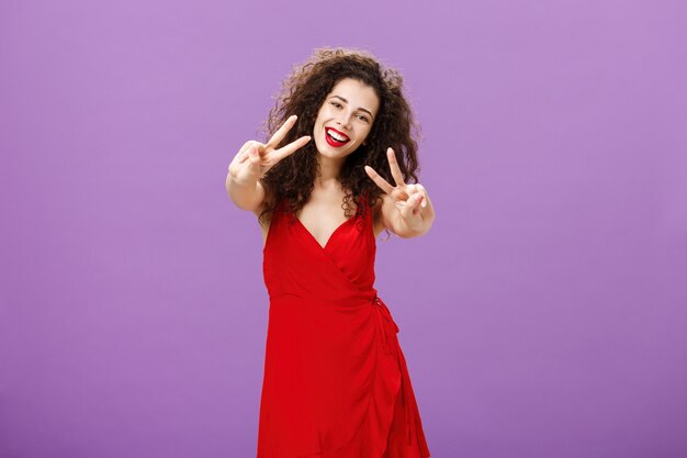 Femme européenne pacifique à l'air sympathique avec une coupe de cheveux bouclée dans une élégante robe rouge montrant la paix ou la vi...