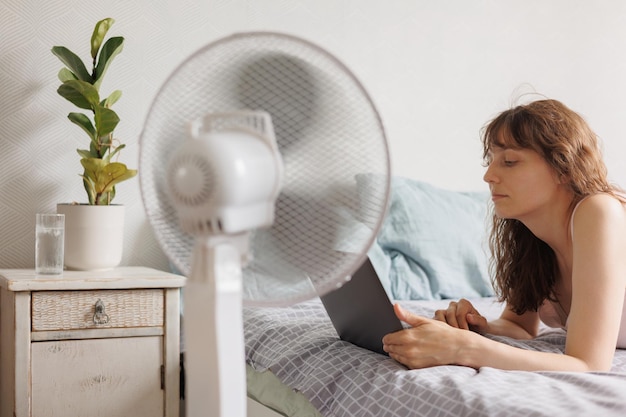 Une femme européenne se refroidit à la maison avec l'aide d'un ventilateur de climatiseur fonctionne avec un ordinateur portable dans le bureau à domicile