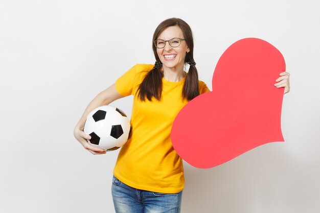 Femme européenne souriante, queues de cheval amusantes, fan de football ou joueur à lunettes, uniforme jaune tenant un ballon de football classique, coeur rouge isolé sur fond blanc. Sport, football, concept de mode de vie sain.