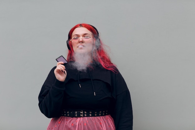 Femme européenne de taille plus dans un casque fumant une cigarette électronique vape Jeune fille positive aux cheveux rouges et roses