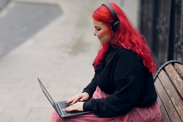 Une femme européenne de taille plus utilise un ordinateur portable alors qu'elle est assise sur un banc en bois à l'extérieur pendant la journée.