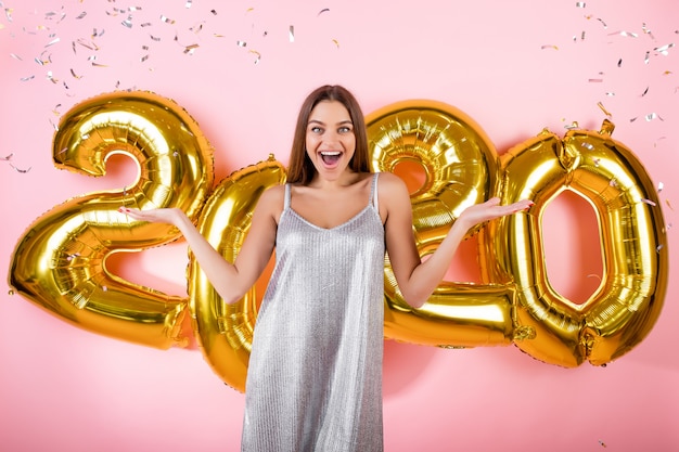 Femme excitée avec des confettis d'argent et ballons dorés du nouvel an 2020 isolés sur rose