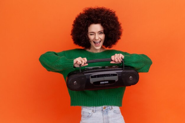 Femme excitée drôle avec une coiffure Afro portant un chandail de style décontracté vert tenant un magnétophone en appuyant sur le bouton écoutant de la musique forte Studio tourné en intérieur isolé sur fond orange