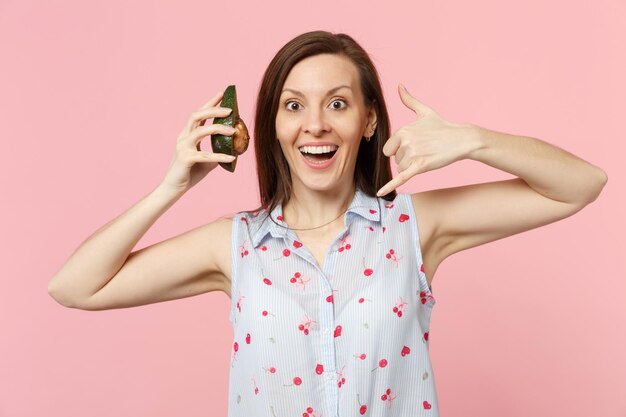 Femme excitée faisant un geste téléphonique comme dit rappelez-moi tenant la moitié des fruits frais d'avocat mûrs isolés sur fond pastel rose. Les gens vivent un style de vie vivant et se détendent. Maquette de l'espace de copie.