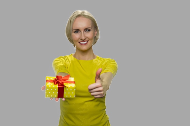 Femme excitée montrant la boîte-cadeau et le pouce vers le haut. Heureuse femme souriante montrant une petite boîte-cadeau et signe d'approbation sur fond gris.
