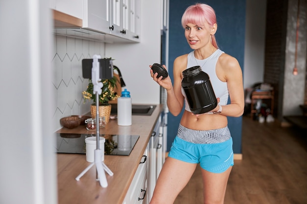 Une femme excitée montre un grand pot de complément alimentaire à la caméra dans la cuisine