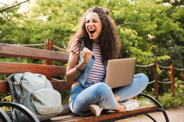 Femme excitée ravie des années 20 avec les cheveux bouclés serrant le poing alors qu'elle était assise sur un banc dans un parc verdoyant et étudie ou travaille sur un ordinateur portable