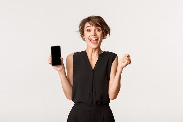 Femme excitée à la recherche de plaisir tout en montrant l'écran du smartphone, souriant amusé, debout blanc.