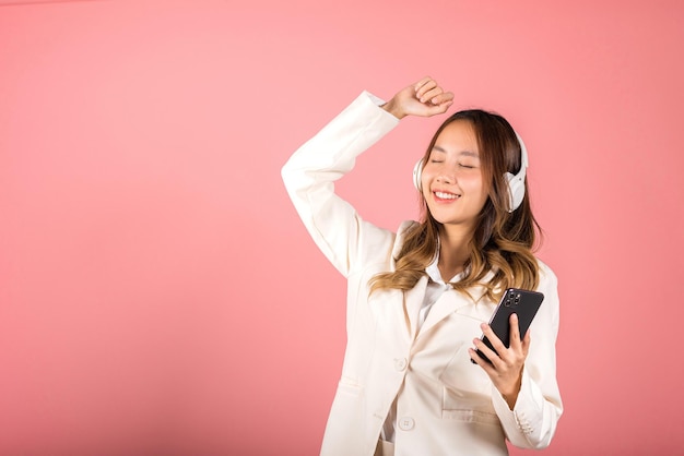 Femme excitée souriante écoutant de la radio musicale dans un casque Bluetooth et levant la main dansant sur un studio de smartphone tourné en arrière-plan blanc isolé, application en ligne de jeune femme asiatique heureuse sur téléphone mobile