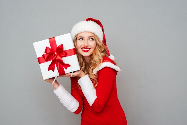 Femme excitée souriante en tenue de père noël rouge tenant un cadeau de noël à son oreille isolée sur la surface grise