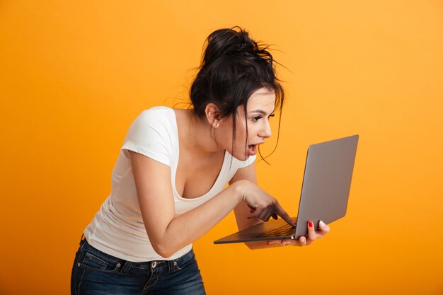 Femme excitée en t-shirt décontracté et jeans en tapant un message texte ou en naviguant sur Internet à l'aide d'un ordinateur portable argenté, sur un mur jaune