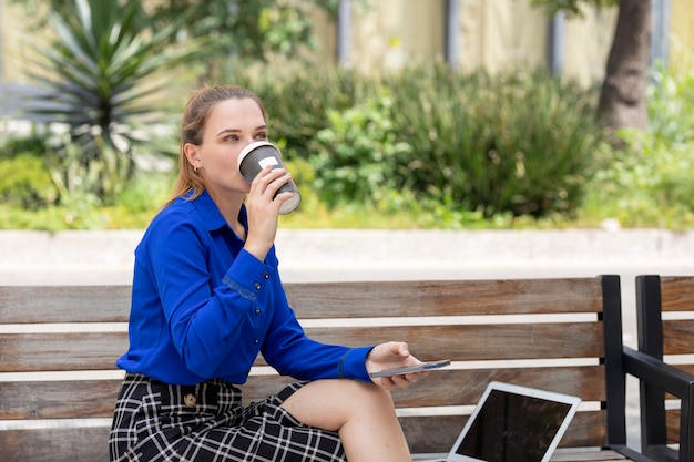 Une femme exécutive boit du café dans un verre tout en travaillant sur son ordinateur portable assis sur un banc de rue