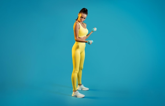 Femme exerçant tenant des haltères fléchissant les biceps ayant un fond bleu d'entraînement