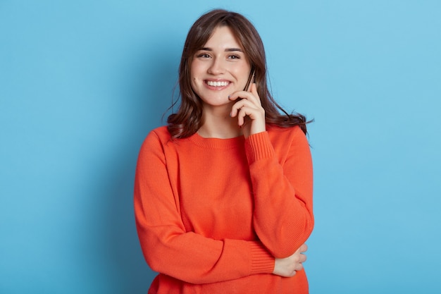 femme avec une expression faciale heureuse posant avec le téléphone près de l'oreille isolée sur le mur bleu.