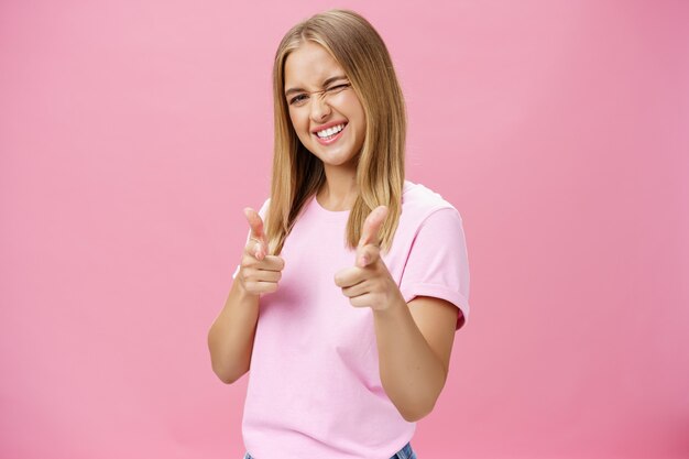 Femme exprimant une attitude positive envers la caméra pointant avec les doigts et un clin d'œil joyeusement sur fond rose.