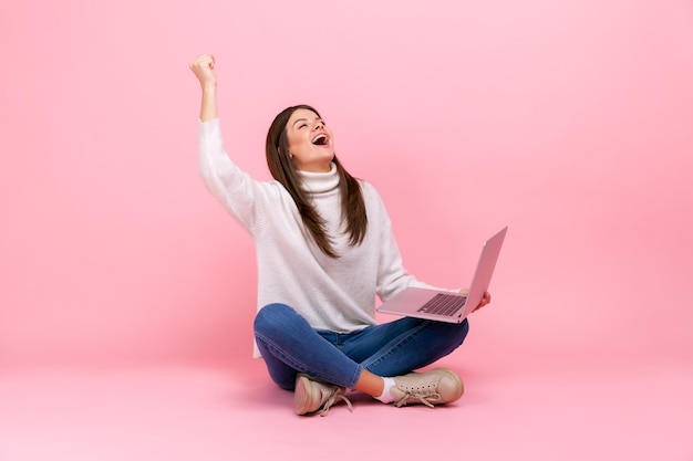 Femme extrêmement heureuse assise sur le sol avec un ordinateur portable criant pour célébrer sa victoire ou son succès portant un pull blanc de style décontracté Prise de vue en studio intérieure isolée sur fond rose