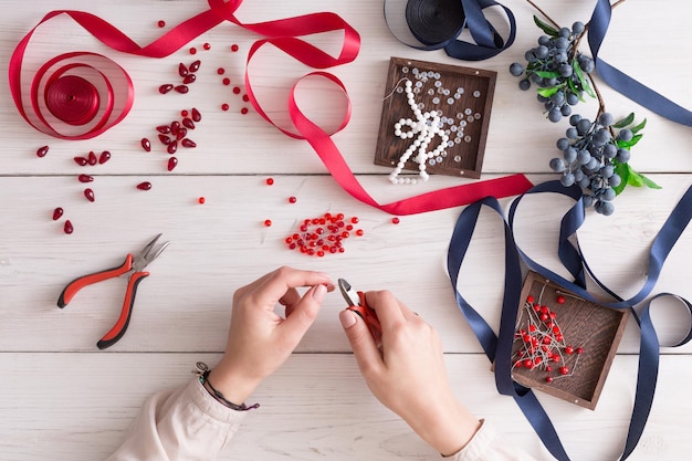 Femme faisant des bijoux, atelier à domicile. Artisan pov, mains féminines créant un accessoire avec des perles et des rubans, vue de dessus. Beauté, créativité, concept d'artisanat