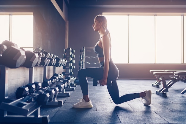 Femme faisant des exercices de fentes avec des haltères dans une salle de sport.