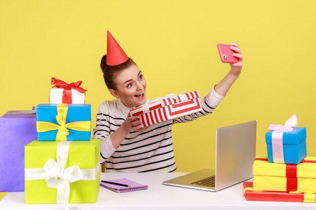 Femme faisant selfie sur smartphone caméra assis sur le lieu de travail avec de nombreux coffrets cadeaux colorés autour