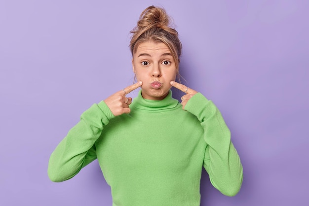 la femme fait la moue et pointe les joues qui soufflent fait la grimace devant la caméra vêtue d'un col roulé vert décontracté isolé sur violet. Concept d'expressions de visage humain