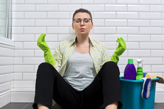 Femme fatiguée assise dans la salle de bain sur le sol et méditant. Femme en salle blanche avec seau de détergent au repos en méditation