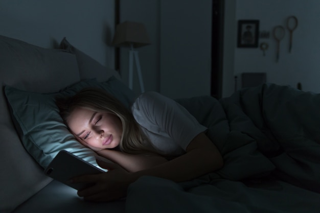 Photo femme fatiguée endormie allongée dans son lit sous la couverture à l'aide de smartphone pendant la nuit, ne peut pas dormir.