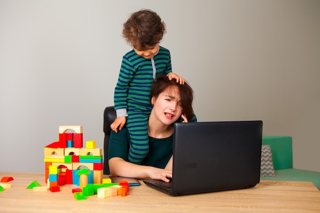 Femme fatiguée avec un enfant sur le cou, assise devant un ordinateur et parlant au téléphone avec l'employeur pendant que l'enfant joue aux cubes et traîne autour d'elle. incapacité de travailler à la maison.