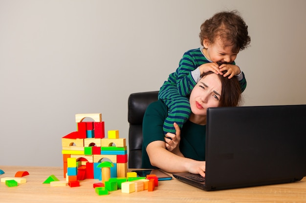 Femme fatiguée avec un enfant sur le cou, assise devant un ordinateur et parlant au téléphone avec l'employeur pendant que l'enfant joue aux cubes et traîne autour d'elle.