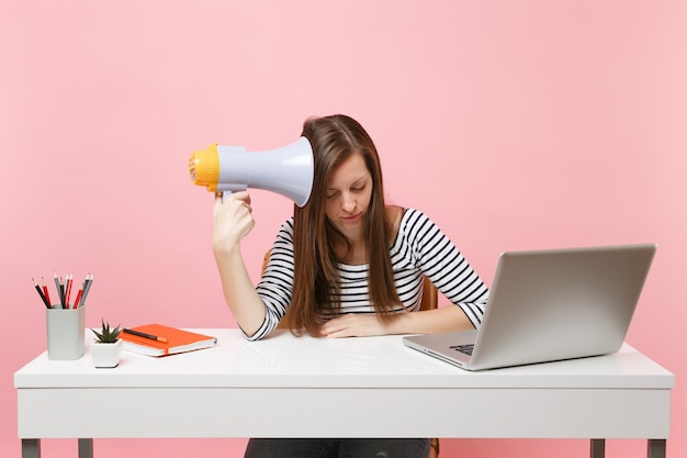 Femme fatiguée épuisée tenant un mégaphone assis, travaillant sur un projet au bureau blanc au bureau avec un ordinateur portable isolé sur fond rose pastel. Concept de carrière d'entreprise de réalisation. Espace de copie.