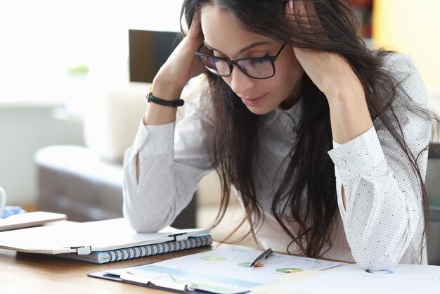 Femme fatiguée avec des lunettes s'asseoir à table et masse sa tête avec ses mains et pense