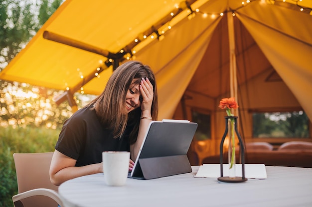 Femme fatiguée pigiste utilisant un ordinateur portable sur une tente de glamping confortable par une journée ensoleillée Tente de camping de luxe pour les vacances d'été en plein air et les vacances Concept de style de vie