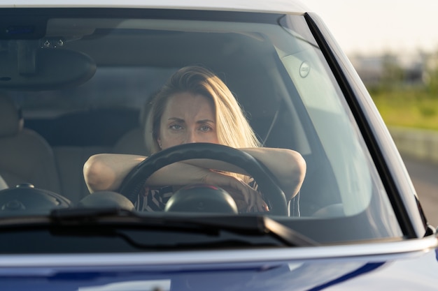 Femme fatiguée réfléchie conduisant une voiture regardant à travers le pare-brise à la caméra en pensant à quelque chose