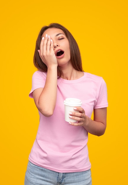 Femme fatiguée avec une tasse de café pour aller bâiller et se frotter le visage sur fond jaune le matin