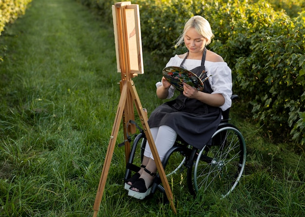 Photo femme en fauteuil roulant à l'extérieur dans la peinture de la nature avec chevalet