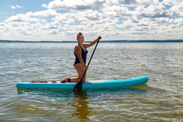 Une femme féministe juive rasée en maillot de bain fermé agenouillée sur une planche SUP avec une rame flotte sur l'eau